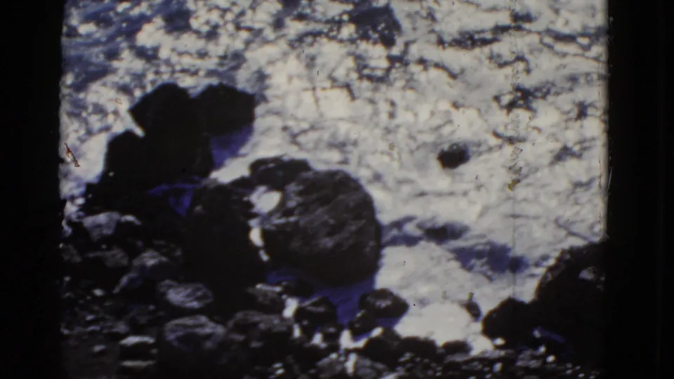 1952 San Francisco coastal frame with a rounded shoreline rock, dark stones, and bright surf filling the background.