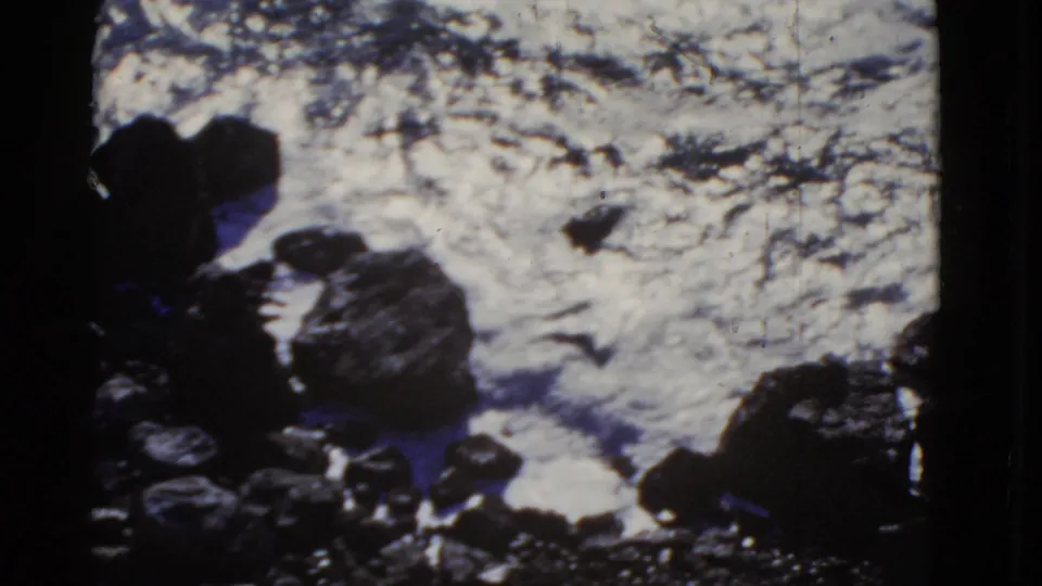1952 San Francisco home-movie frame showing white ocean water breaking against dark coastal boulders from above.