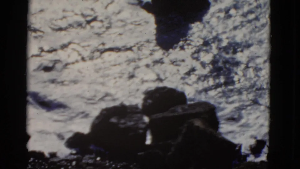 1952 San Francisco 8mm frame of swirling ocean foam beside large boulders on a rocky coast.