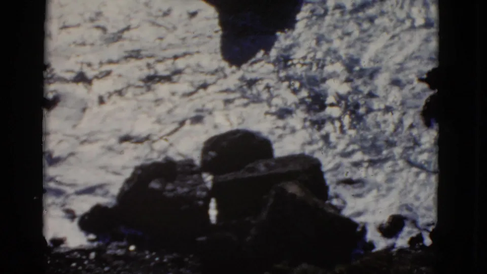 1952 San Francisco home-movie frame showing dark shoreline rocks with foamy surf and a cropped film-frame edge.