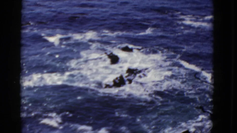 A 1952 San Francisco coastal frame shows white surf spreading over dark rocks in deep blue water.