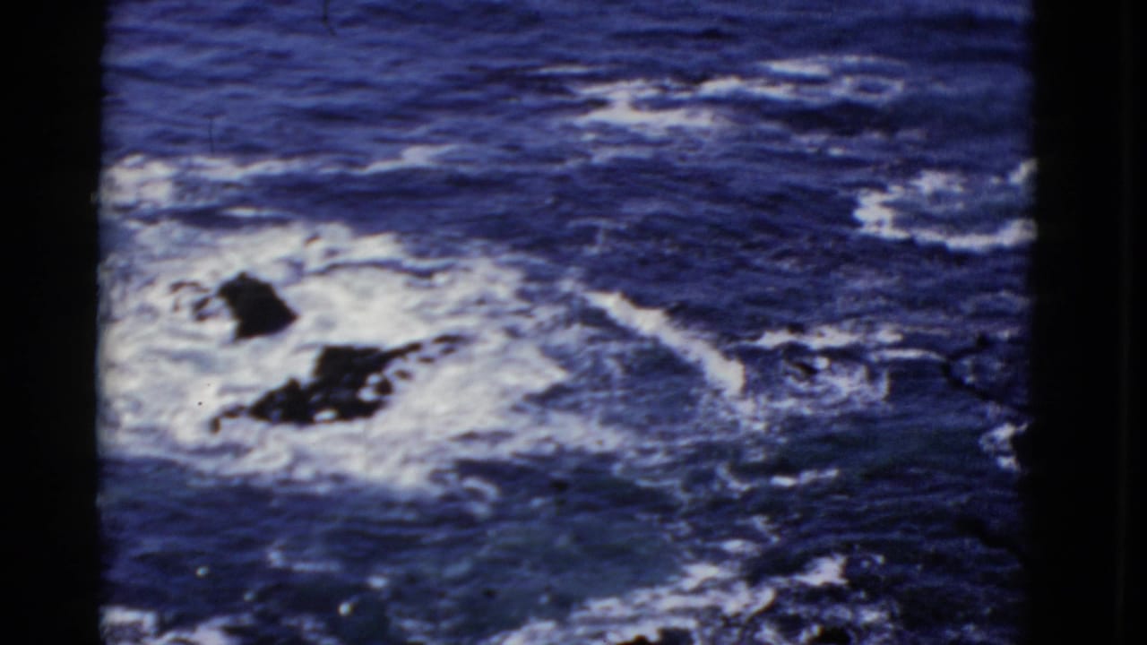 1952 San Francisco 8mm frame showing blue waves and white foam crashing around dark coastal rocks.
