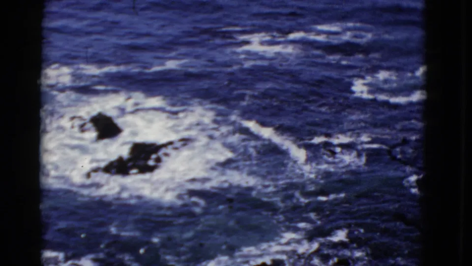 1952 San Francisco 8mm frame showing blue waves and white foam crashing around dark coastal rocks.
