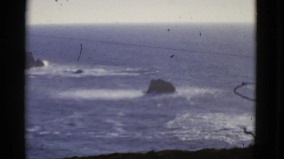 1952 home-movie frame of the San Francisco coast with distant horizon, offshore rocks, and pale surf lines.