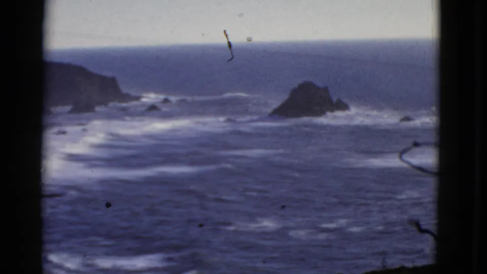 1952 8mm coastal frame from San Francisco showing a dark headland, rocky outcrop, blue surf, and soft home-movie focus.