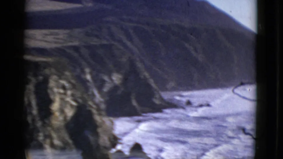 1952 San Francisco coastal home-movie frame showing waves along a rocky shore beneath a shadowed cliff.