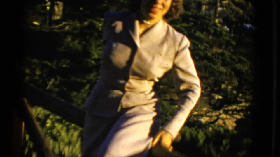 1952 San Francisco frame showing an adult woman in a light-colored suit smiling outdoors beside dense green foliage.