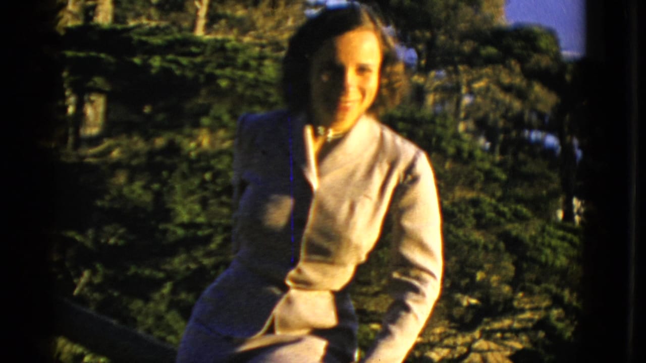 Woman in a light suit sits outdoors in front of trees and distant hills.