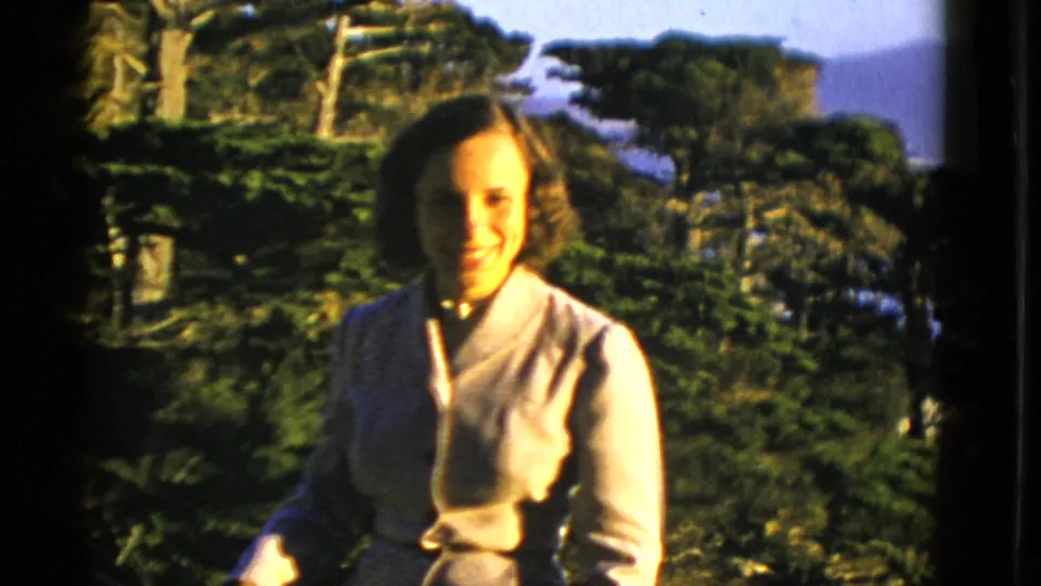 A woman in a pale suit smiles toward the camera, backed by greenery and distant San Francisco hillside or bay scenery.