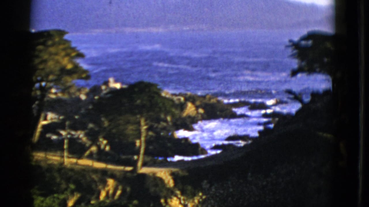Coastal overlook with surf, rocks, trees, road, ocean, and distant hills.