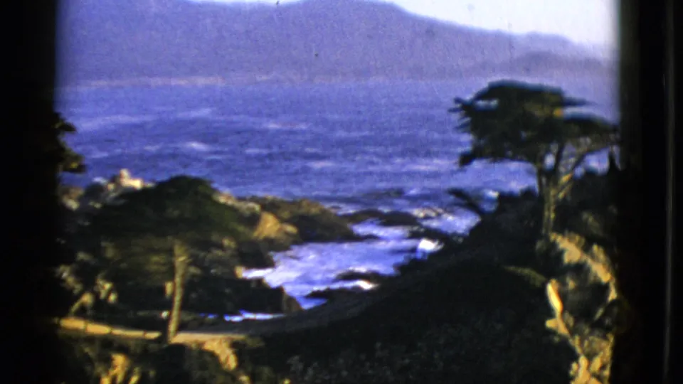 1952 San Francisco-area coastal frame showing white surf in a rocky cove, dark windblown trees on the headland, and hills across the water.
