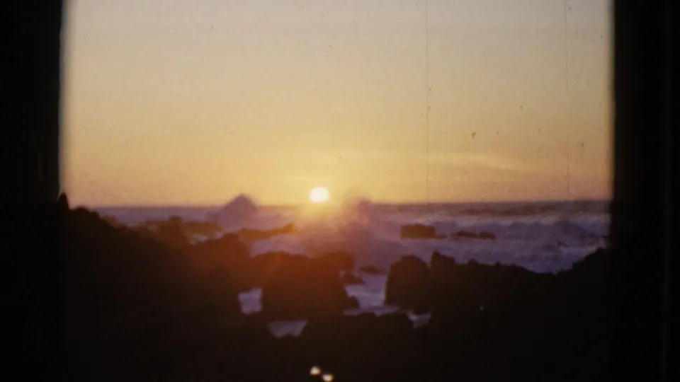 1952 8mm sunset view over the ocean, with the sun near the horizon and dark coastal rocks silhouetted in front.