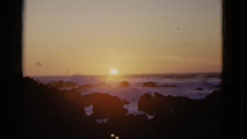 1952 San Francisco 8mm frame showing a low sun over the ocean, silhouetted coastal rocks, and a dark vertical window frame at the edge of the view.