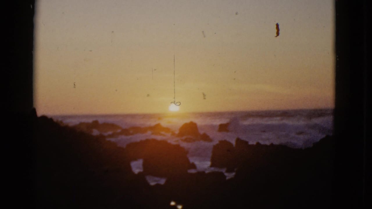 1952 San Francisco coastal sunset view with the sun close to the horizon, ocean waves around dark rocks, and vertical window framing visible in the foreground.