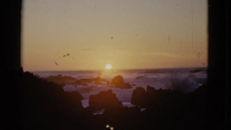 1952 San Francisco 8mm still with a warm sunset over the ocean, dark shoreline rocks in the foreground, and visible dust and scratches in the film scan.