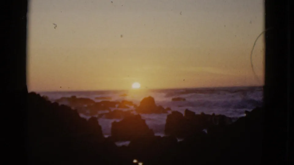 1952 San Francisco home-movie frame showing the sun over a dim ocean horizon, waves breaking around shoreline rocks, and a dark window frame shaping the view.