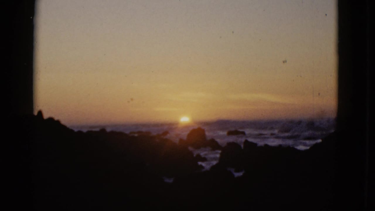 1952 8mm frame of a warm sunset at the ocean horizon, with dark coastal rocks and choppy surf silhouetted in the foreground.