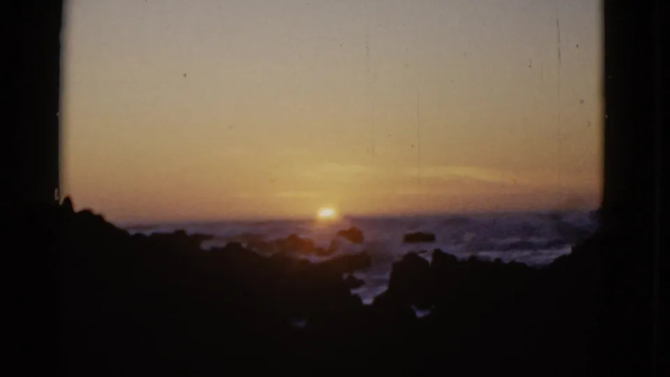 1952 San Francisco coastal home-movie frame with the sun just above the horizon and waves washing through dark rock formations.
