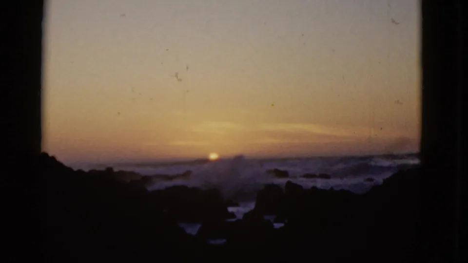 1952 8mm frame showing the sun nearly at the horizon as surf breaks across a rocky California shoreline under a fading evening sky.