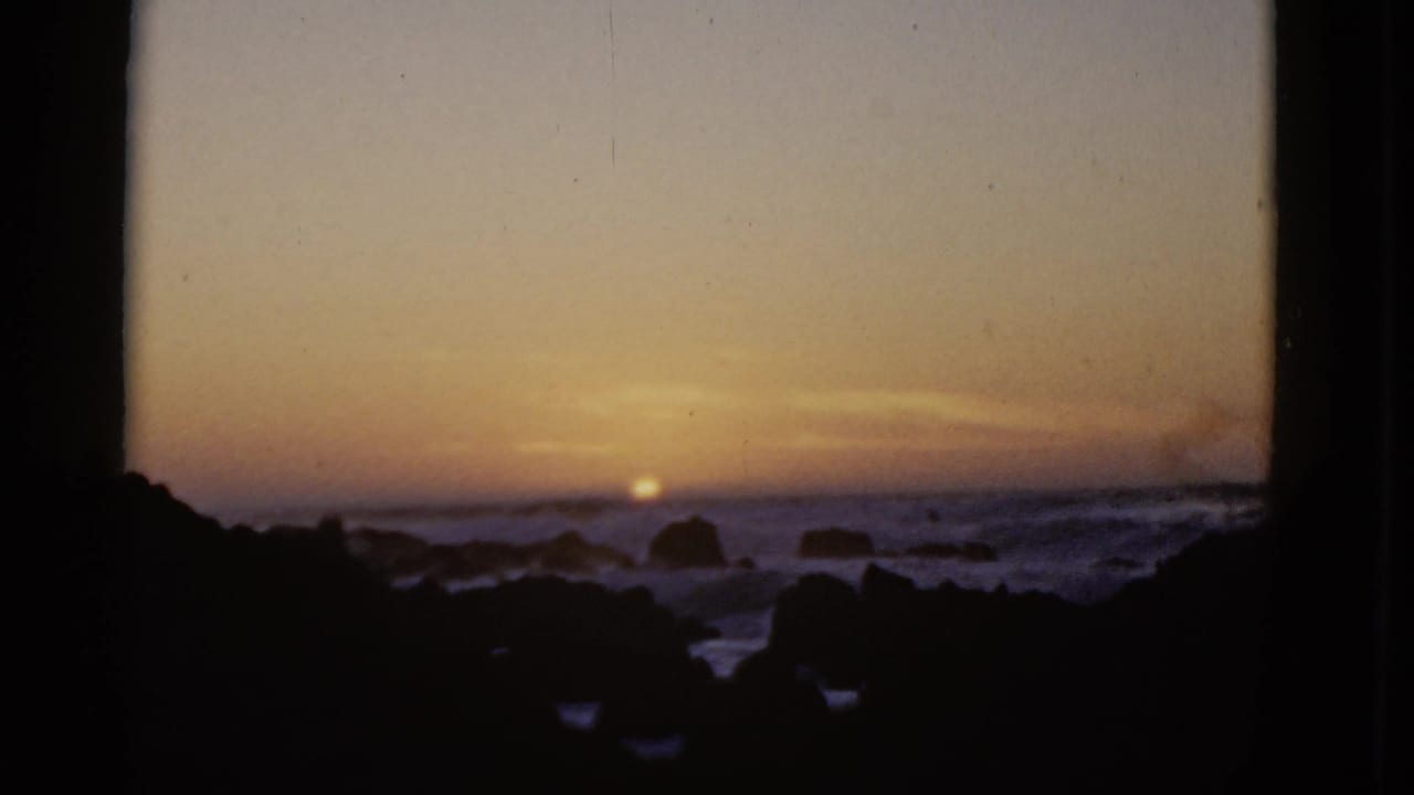 1952 8mm home-movie frame of a low sunset over a rocky San Francisco coastline, with surf and dark foreground rocks.