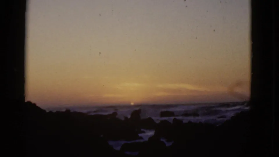 End frame of a 1952 8mm San Francisco coastal sunset, with a small bright sun, surf, and silhouetted rocks.