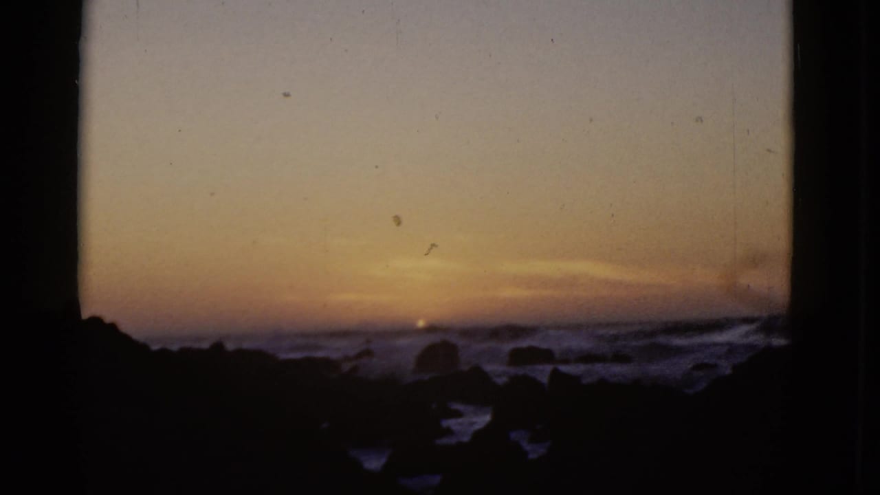1952 8mm view of a warm sunset over San Francisco Bay with rocky shoreline silhouettes and low waves in the foreground.