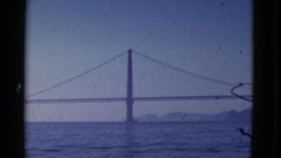 Soft 8mm view of the Golden Gate Bridge spanning San Francisco Bay, with water in the foreground and a muted blue sky behind it.