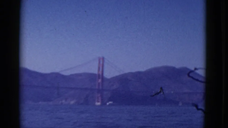 1952 San Francisco 8mm frame showing the Golden Gate Bridge tower and cables above rippled water and distant hills.