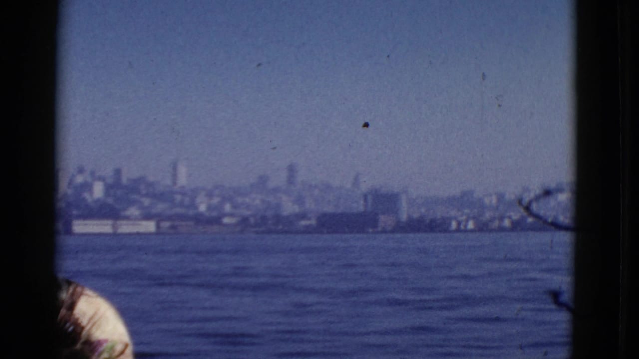 1952 San Francisco bay view from a boat, with hazy shoreline buildings in the distance and part of a passenger at lower left.