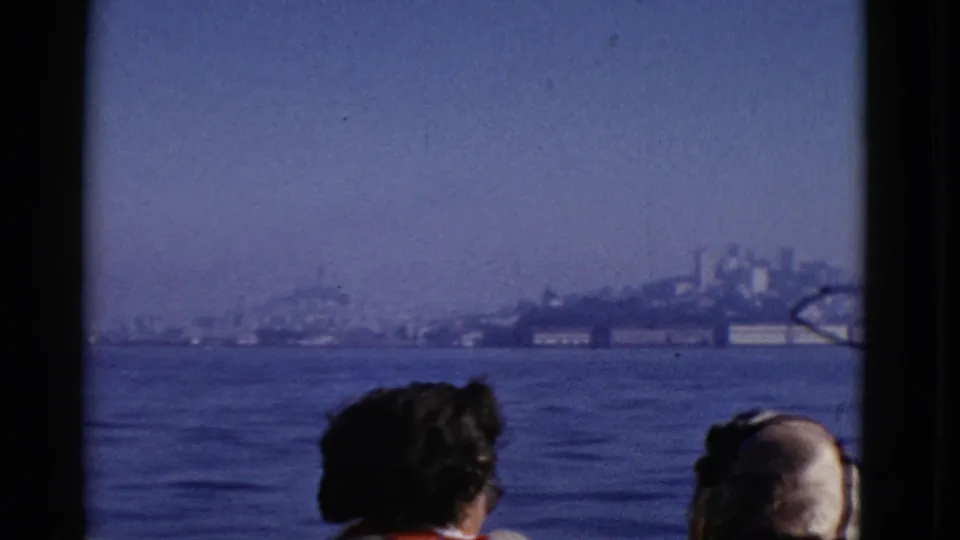 Boat passengers seen from behind in the foreground as the hazy San Francisco shoreline recedes across the bay.