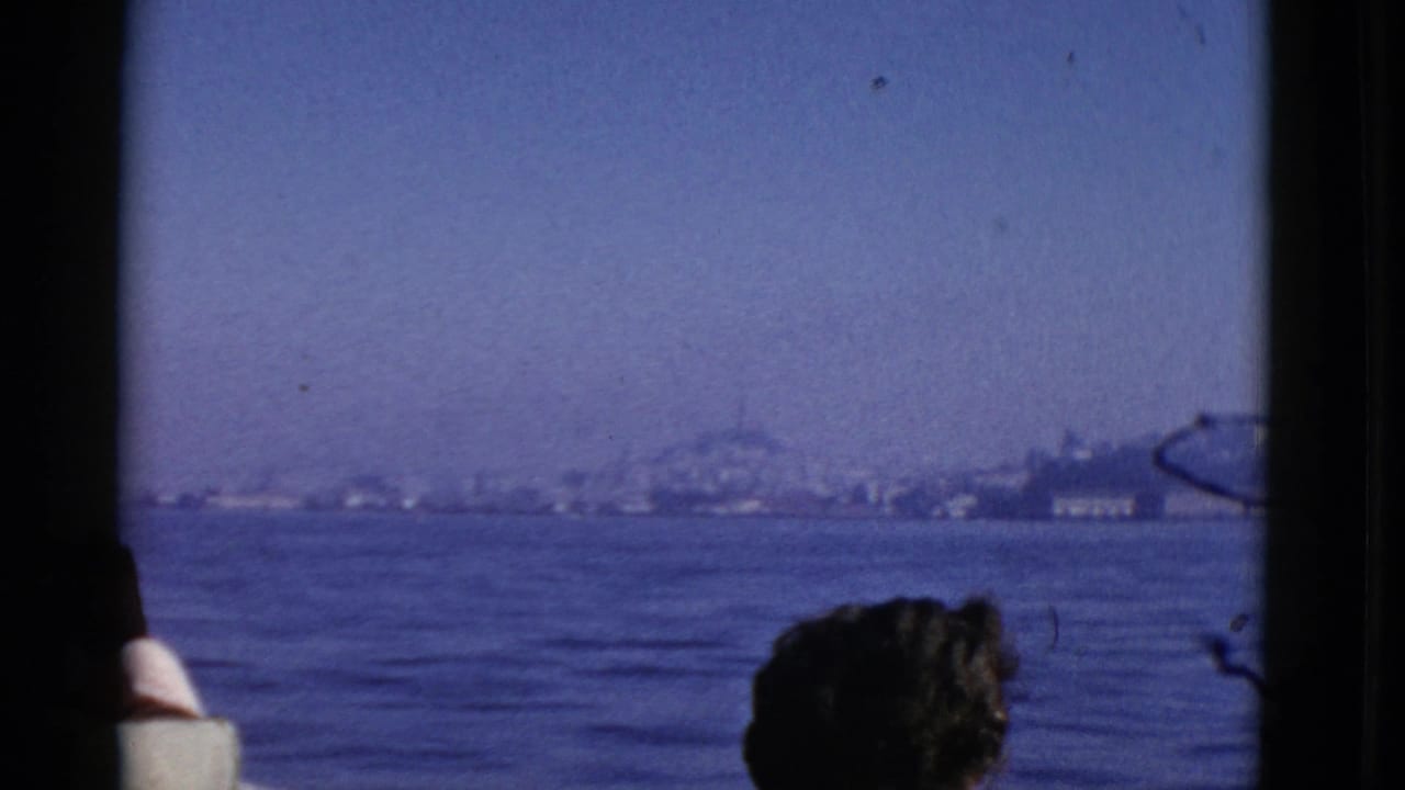 A seated passenger is framed by a boat window with bay water and shoreline beyond.