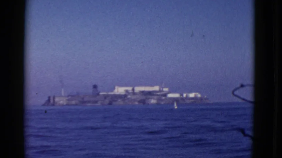 1952 8mm frame of Alcatraz Island seen from a boat on San Francisco Bay, with water in the foreground and haze over the horizon.