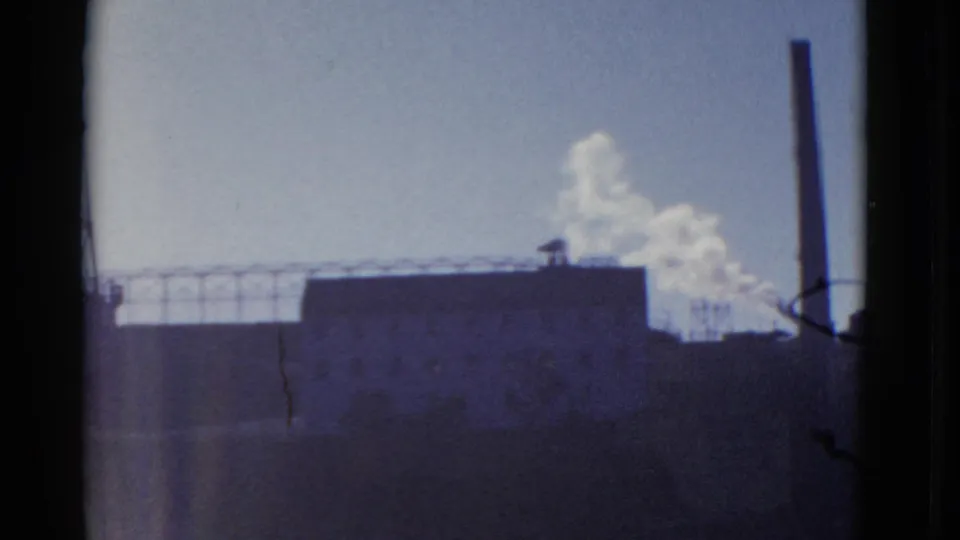 A close 1952 8mm view shows a dark industrial facility, high stack, railings, barbed wire, and white smoke plume.
