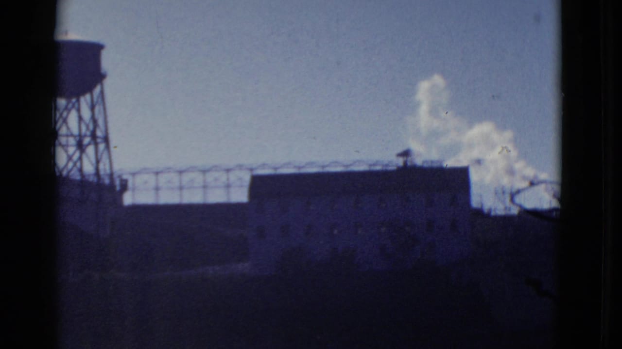 1952 San Francisco industrial exterior with shadowed rooftops, a partial water tower, and white smoke or steam rising near the skyline.