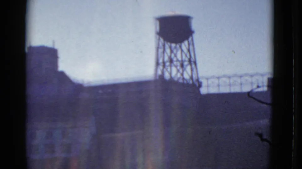 1952 San Francisco industrial exterior showing a water tower, large dark building shapes, and soft vertical light flare on 8mm film.