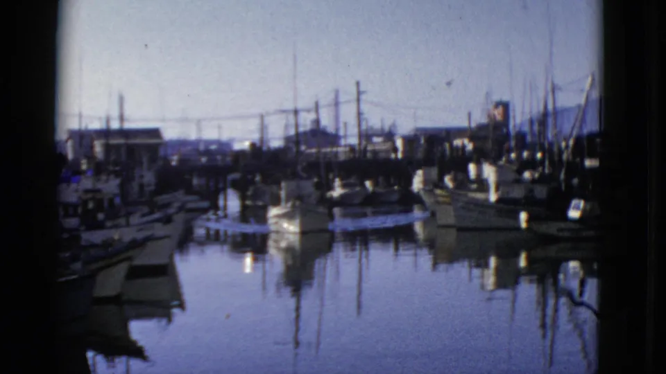Blue-toned 8mm frame of a 1952 San Francisco marina, with a small boat near the dock and masts mirrored in still water.