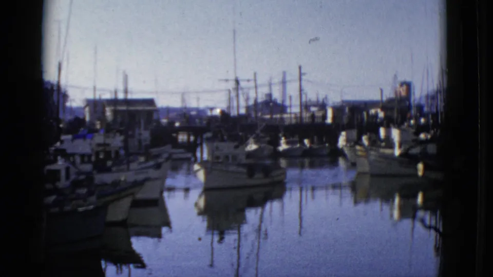 1952 San Francisco waterfront frame showing a small boat near docked vessels, with pilings, masts, and calm reflective water.