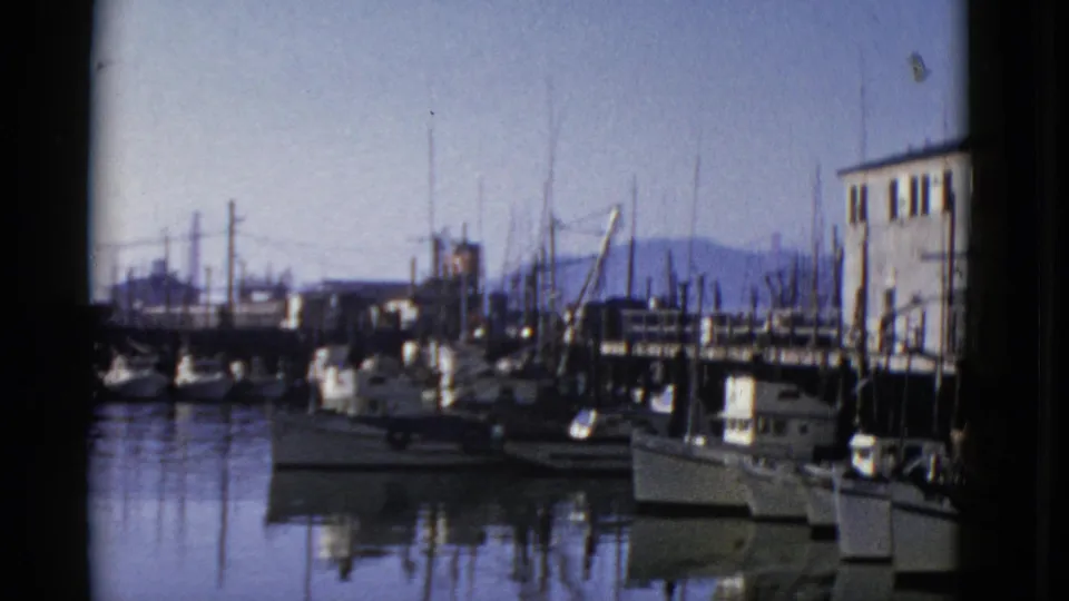 1952 San Francisco dockside view with moored sailboats, a waterfront building at right, calm water, and hills on the horizon.