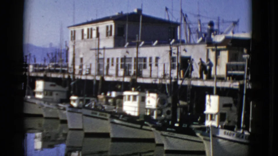 1952 San Francisco harbor frame with docked fishing boats, a light pier building, and a few people visible on the dock.