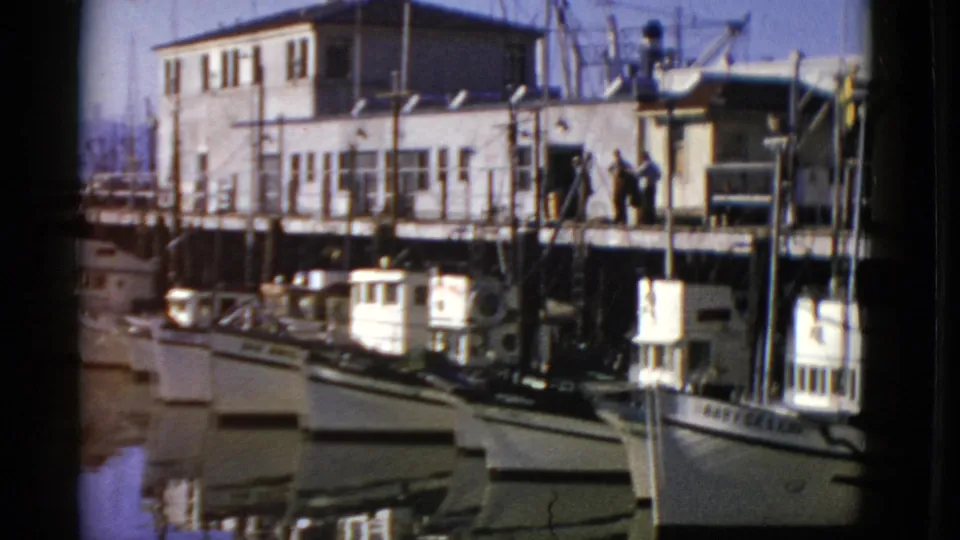 1952 San Francisco harbor view with small fishing boats moored beneath a white pier building and people on the walkway.