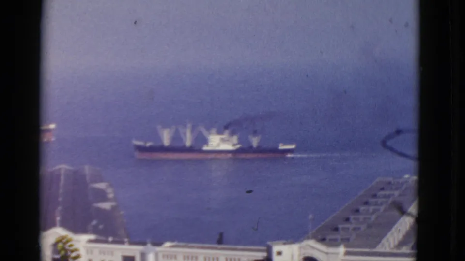 A distant cargo ship moves or sits on the bay beyond a San Francisco waterfront foreground, softened by 8mm film haze.