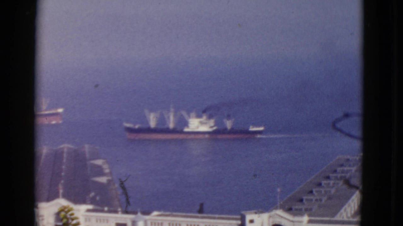 1952 San Francisco harbor view with large working ships crossing the water beyond docks, seen in soft 8mm color.