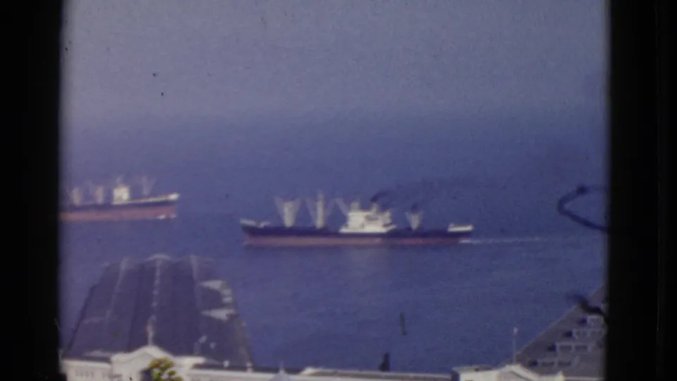 Soft 8mm frame of San Francisco harbor in 1952, with two large ships passing behind dock roofs and low port buildings.