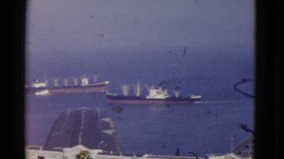 1952 San Francisco Bay footage frame with a large ship and smaller vessel passing beyond sloped foreground roofs.