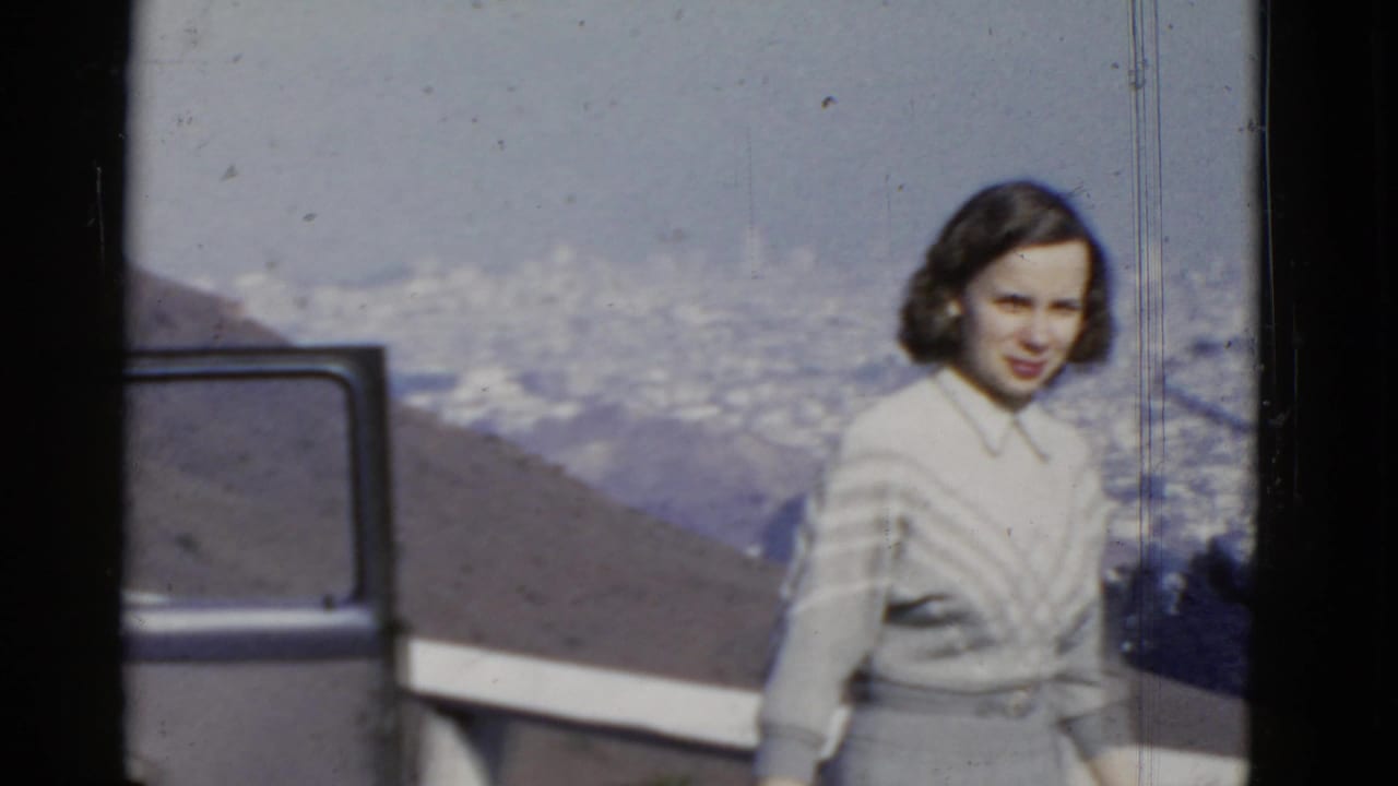 1952 San Francisco home-movie frame showing a woman near an overlook, with a partial vehicle door and hazy cityscape in the background.