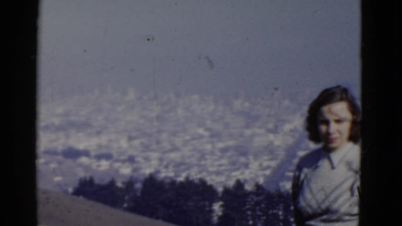 Adult woman poses at a high San Francisco vantage point in 1952, with trees and a hazy cityscape behind her.