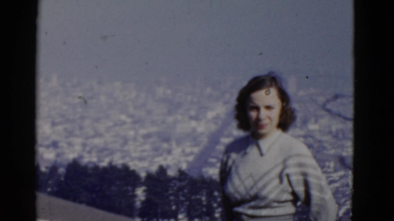 Adult woman faces the camera with a hazy cityscape and dark trees behind her.