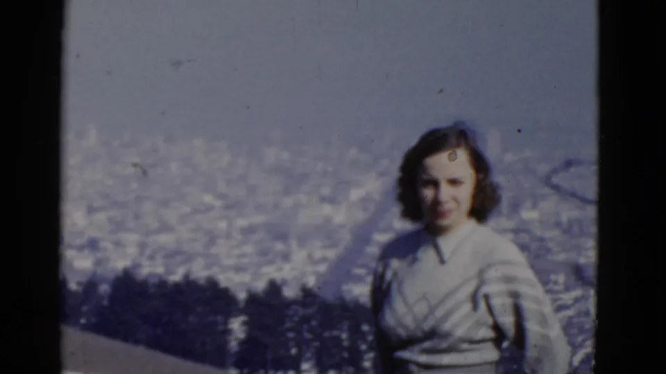 Adult woman in a light sweater faces the camera at a San Francisco overlook, with the distant city softened by haze.