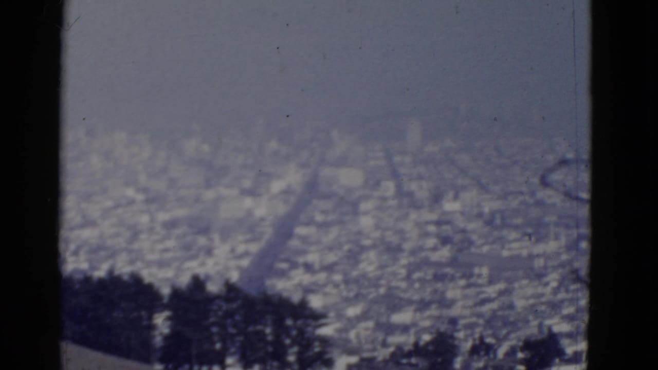 1952 8mm view of San Francisco from an elevated position, showing a hazy city grid beyond silhouetted foreground trees.