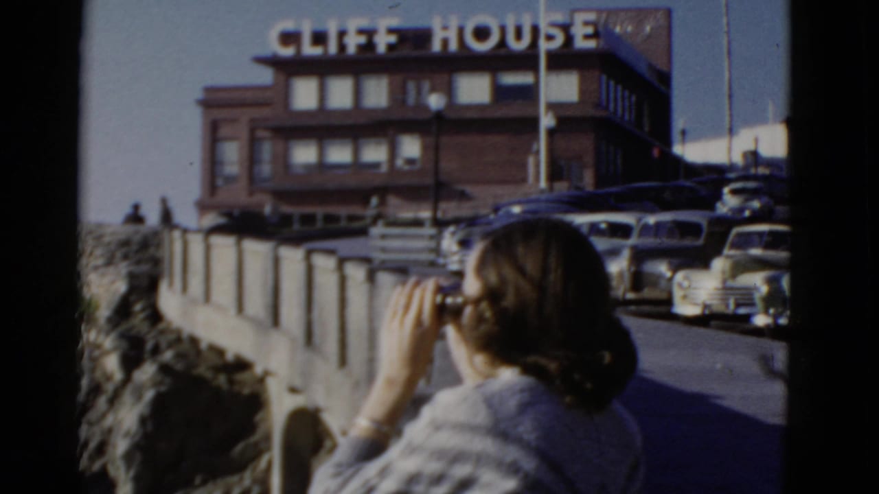 Woman holds binoculars near the Cliff House and parked cars.